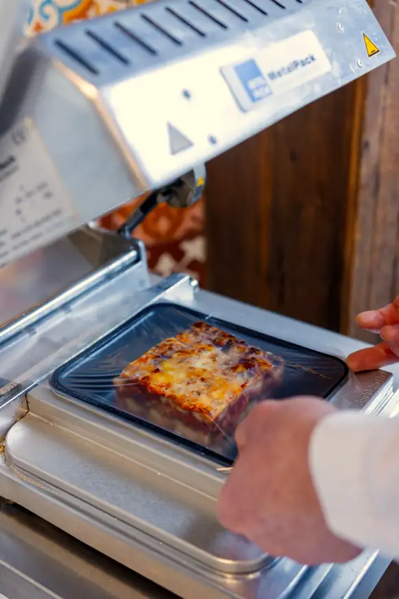 Een huisgemaakte lasagna in een maaltijdbakje die geseald word.